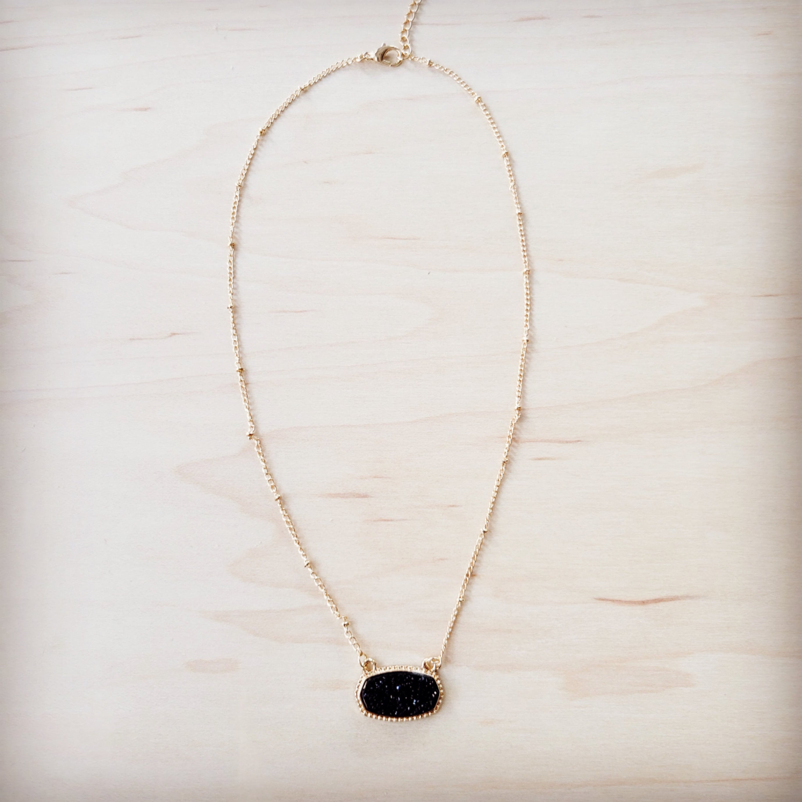 A gold-tone chain necklace with small metal knots and an oval black druzy pendant on a light wood background.