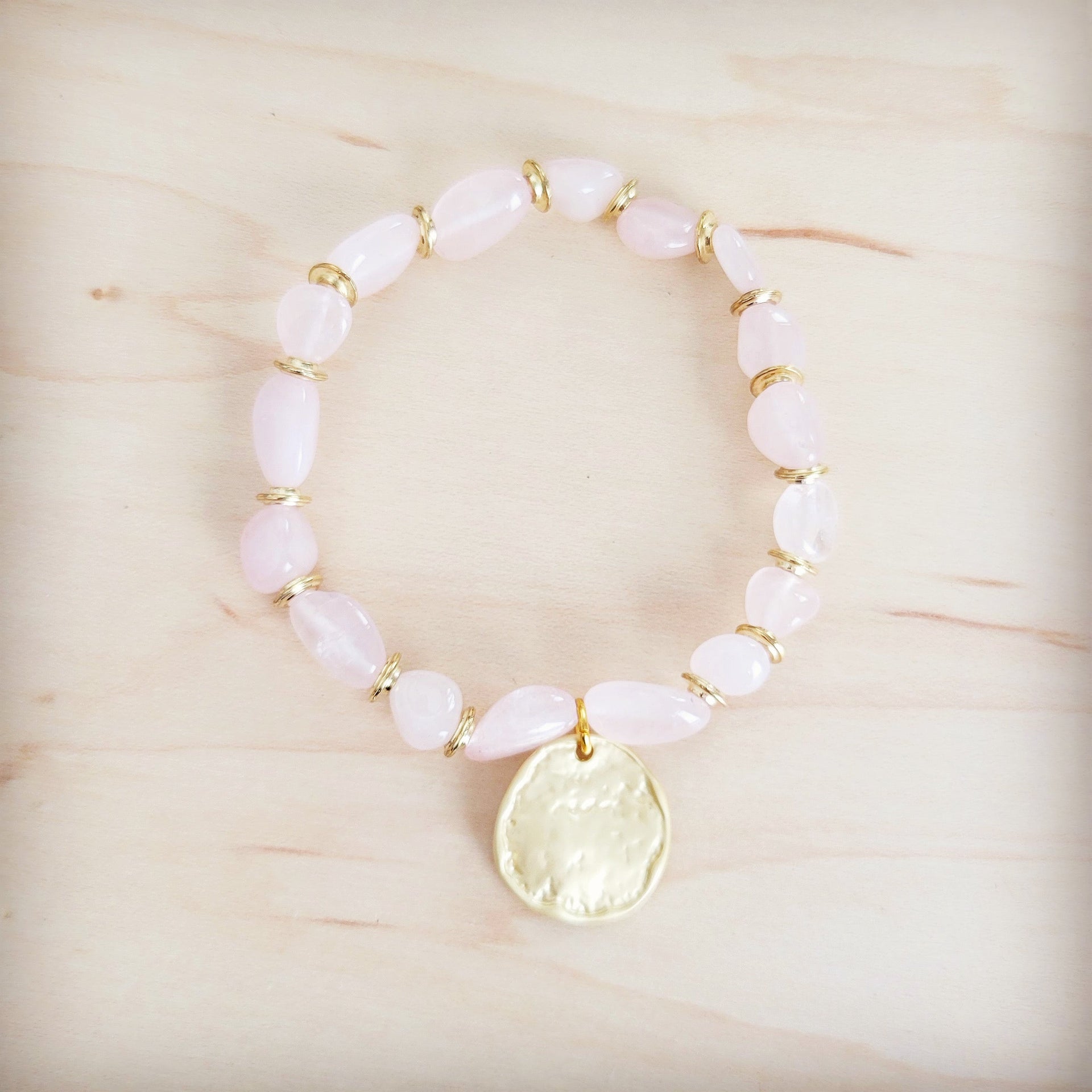 Rose quartz beaded stretch bracelet with a gold-tone coin charm on a light wooden background.