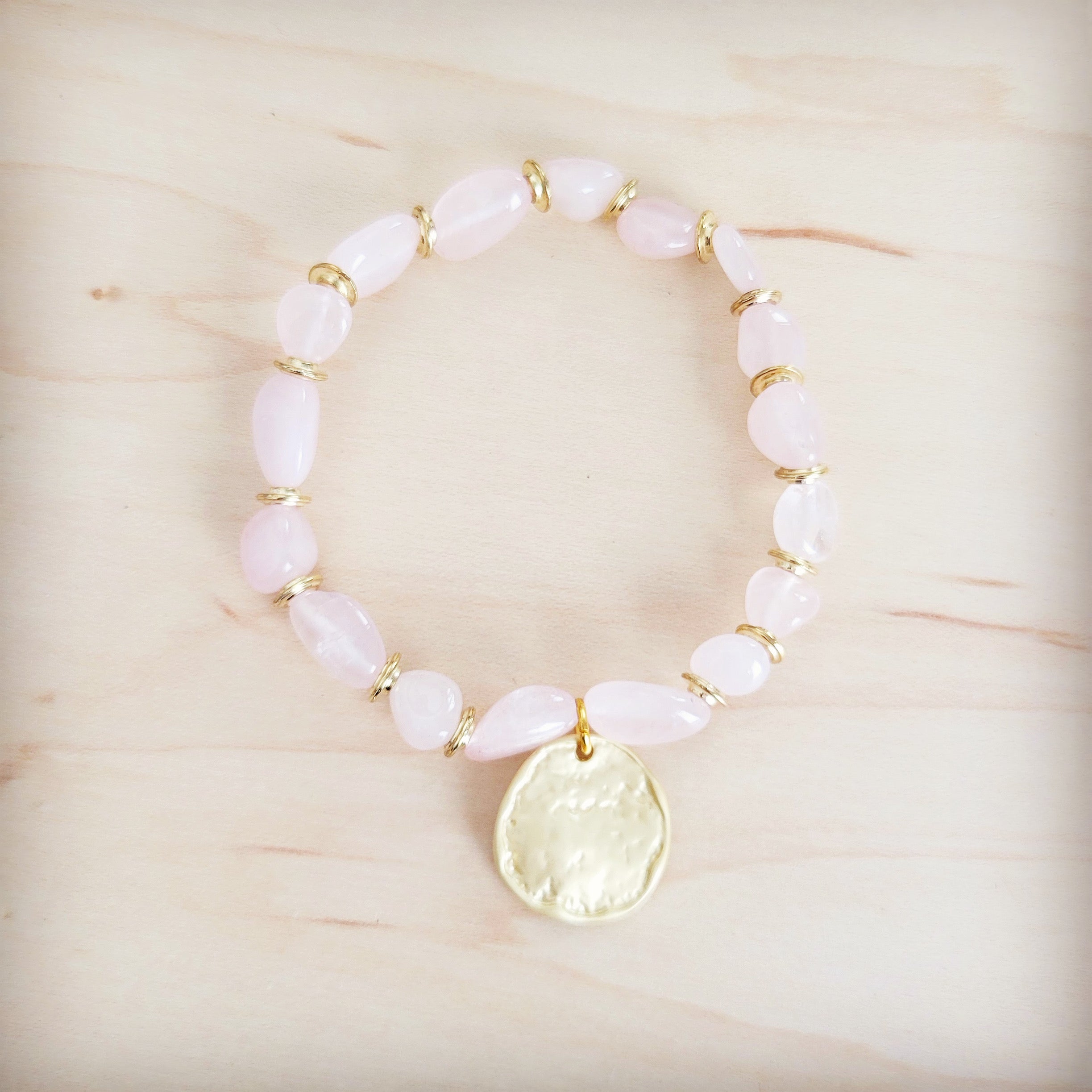 Rose quartz beaded stretch bracelet with a gold-tone coin charm on a light wooden background.