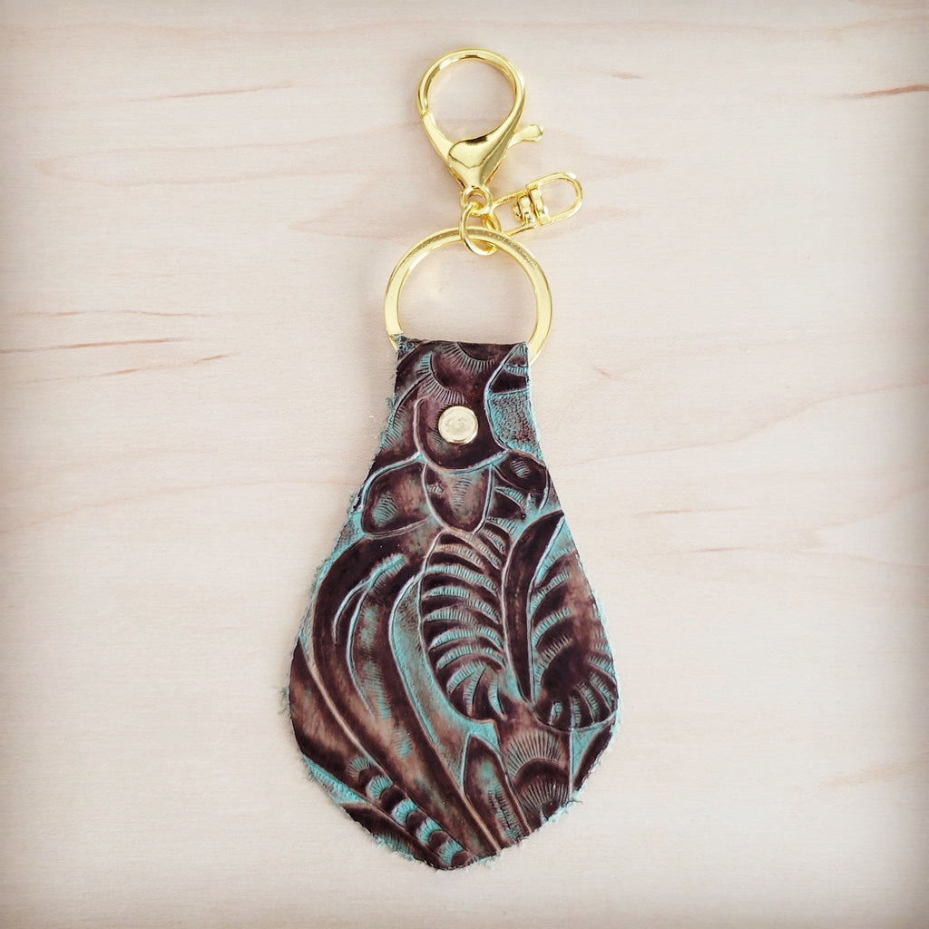 Embossed leather key chain with a turquoise and brown floral pattern and gold-tone metal hardware on a light wood background.