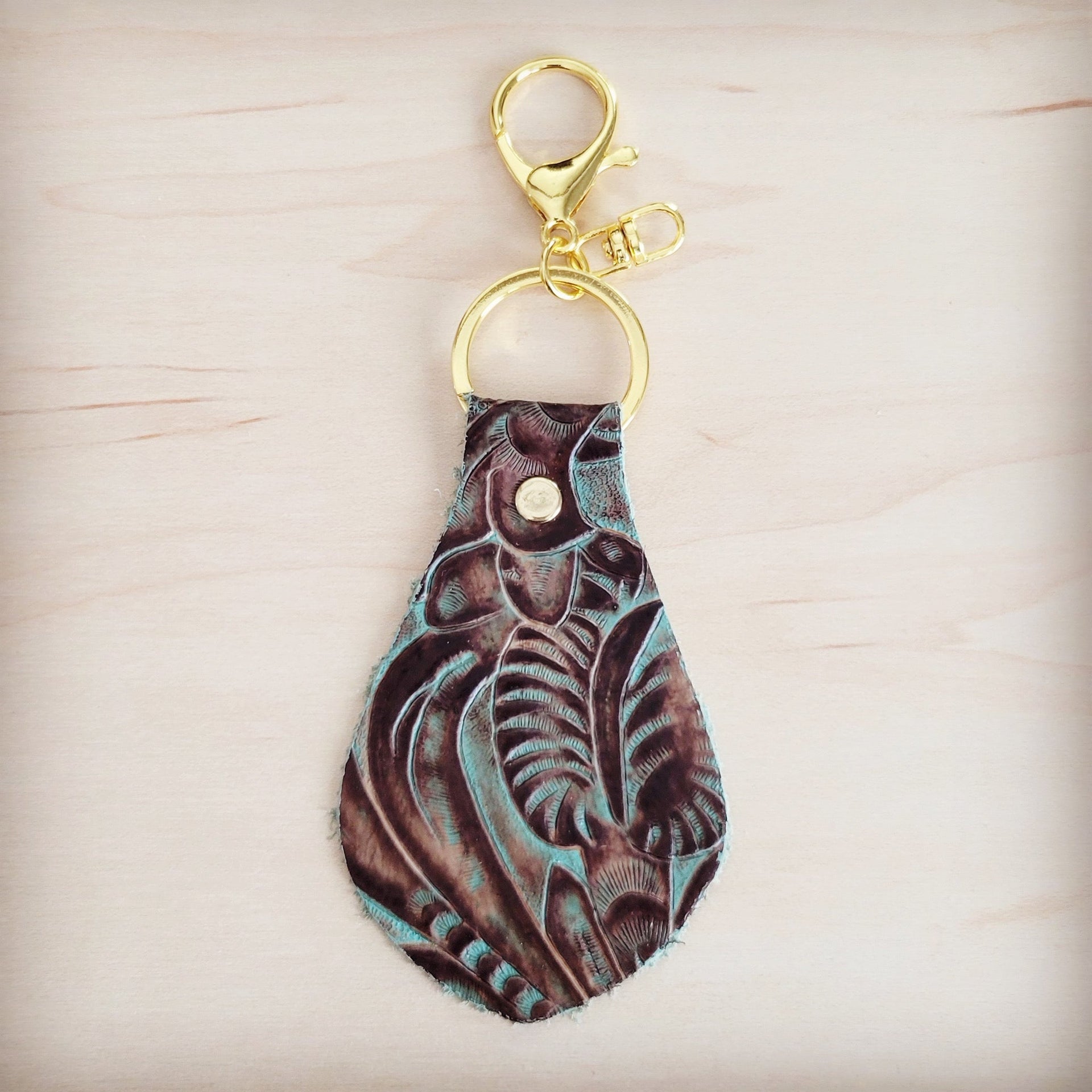 Embossed leather key chain with a turquoise and brown floral pattern and gold-tone metal hardware on a light wood background.