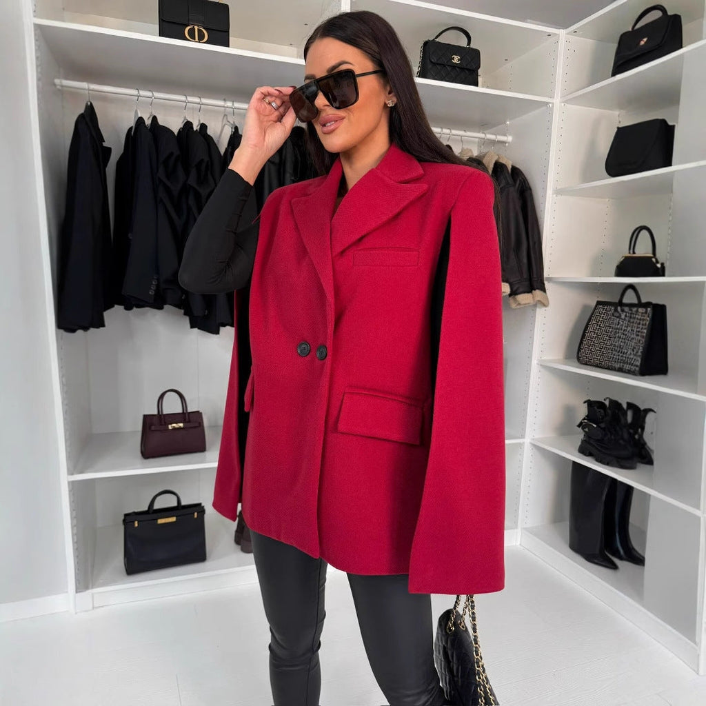A woman wearing a red cape-style blazer with notched lapels over a black top and trousers, standing in a walk-in closet.