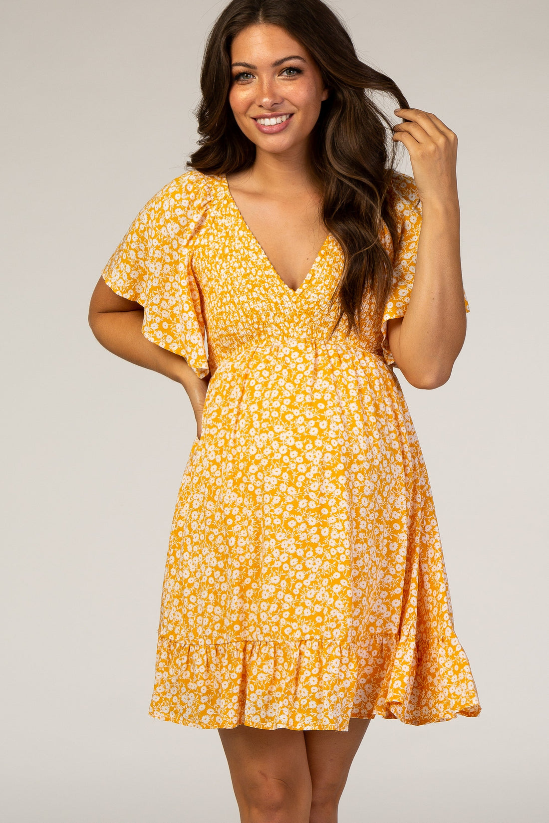 A pregnant woman wearing a bright yellow floral print mini dress with smocked bust and ruffle hem, standing against a neutral studio background.