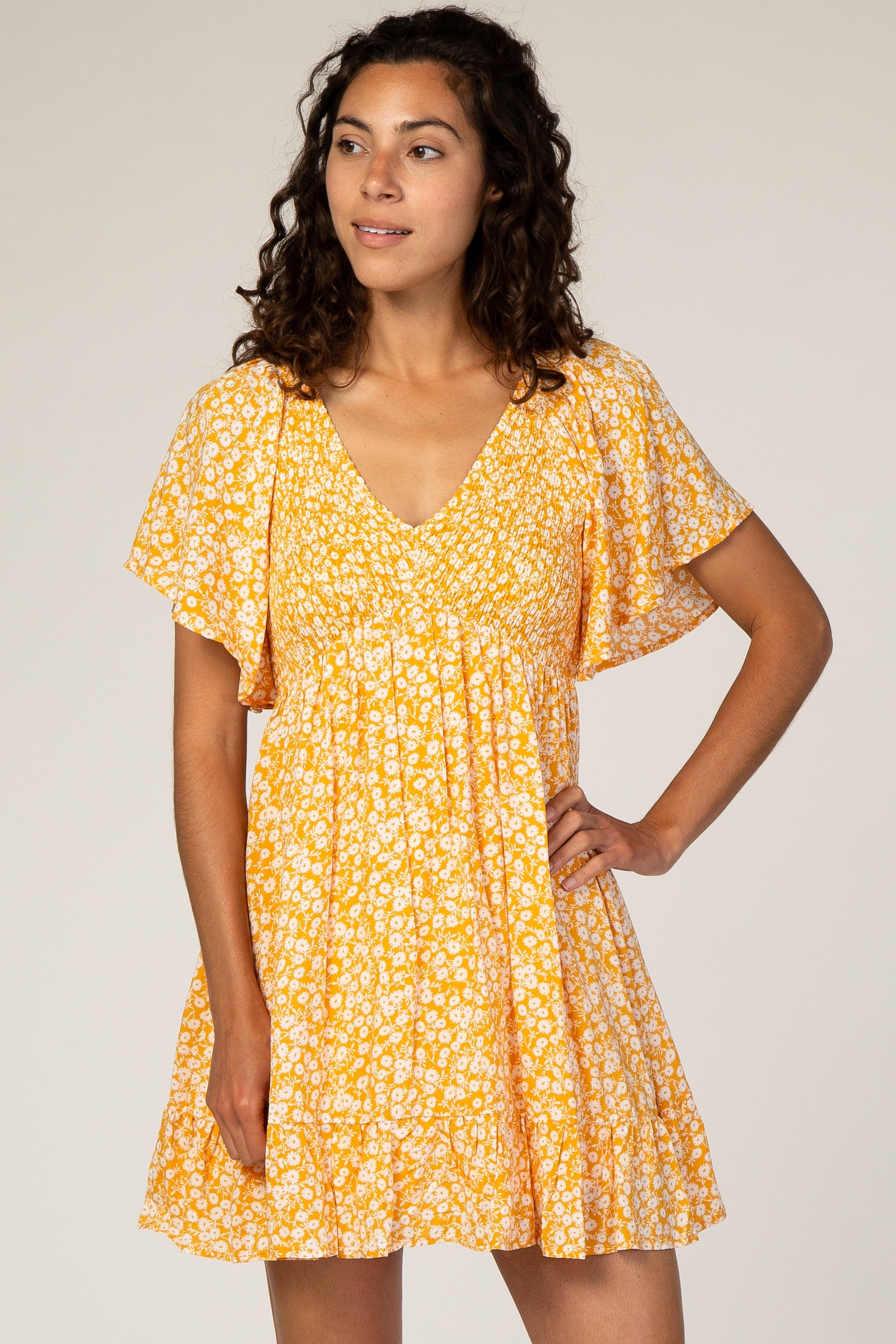 A woman wearing a yellow floral print short dress with smocked bust and ruffle details against a neutral background.