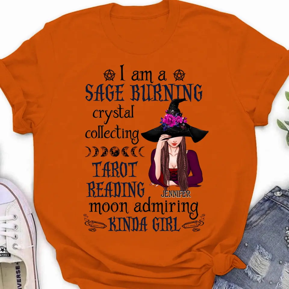 A flat lay of a witch-themed graphic t-shirt featuring sage, crystals, and tarot imagery on an orange and grey background.