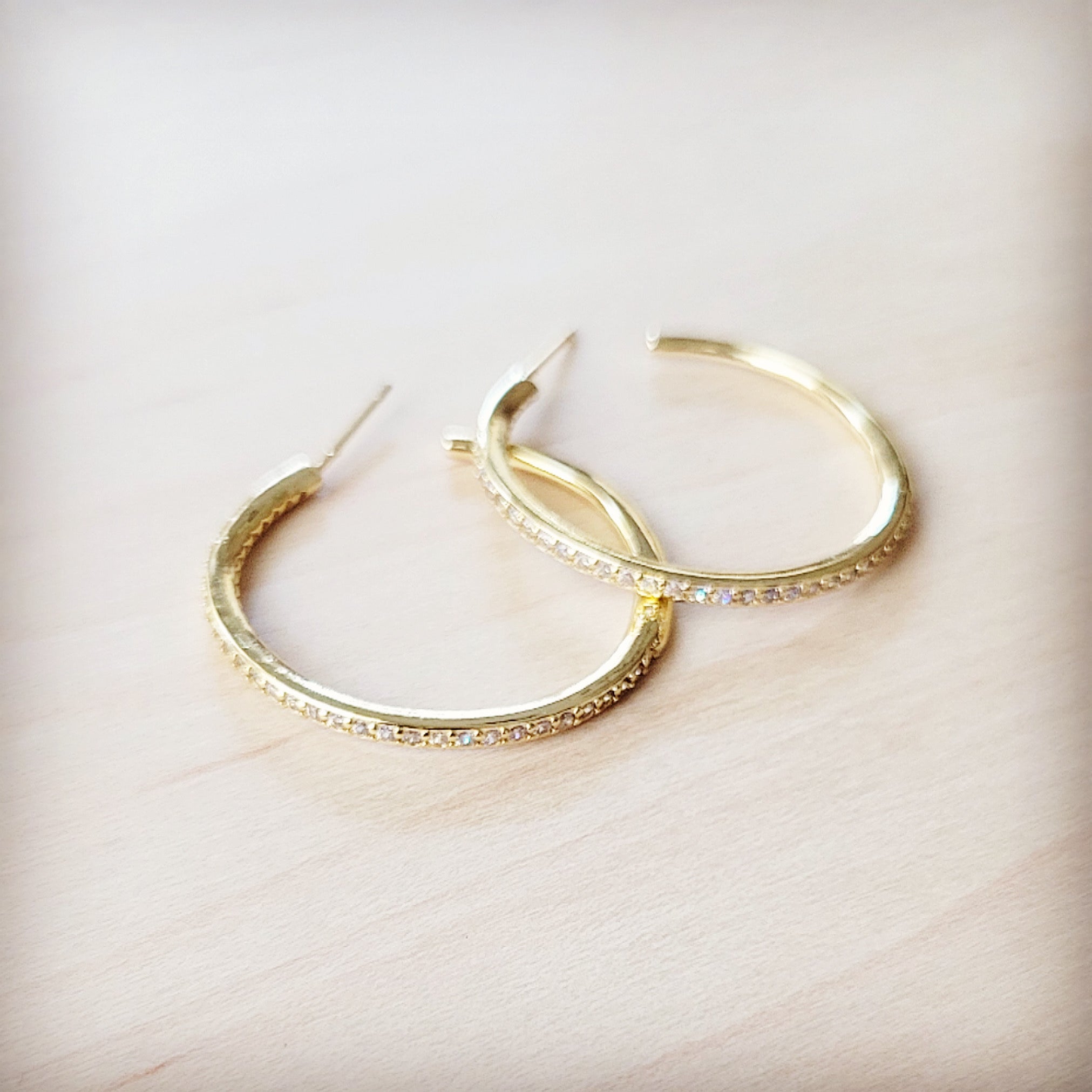 A pair of gold-tone hoop earrings with delicate pave-style crystal inlay resting on a light wooden surface.