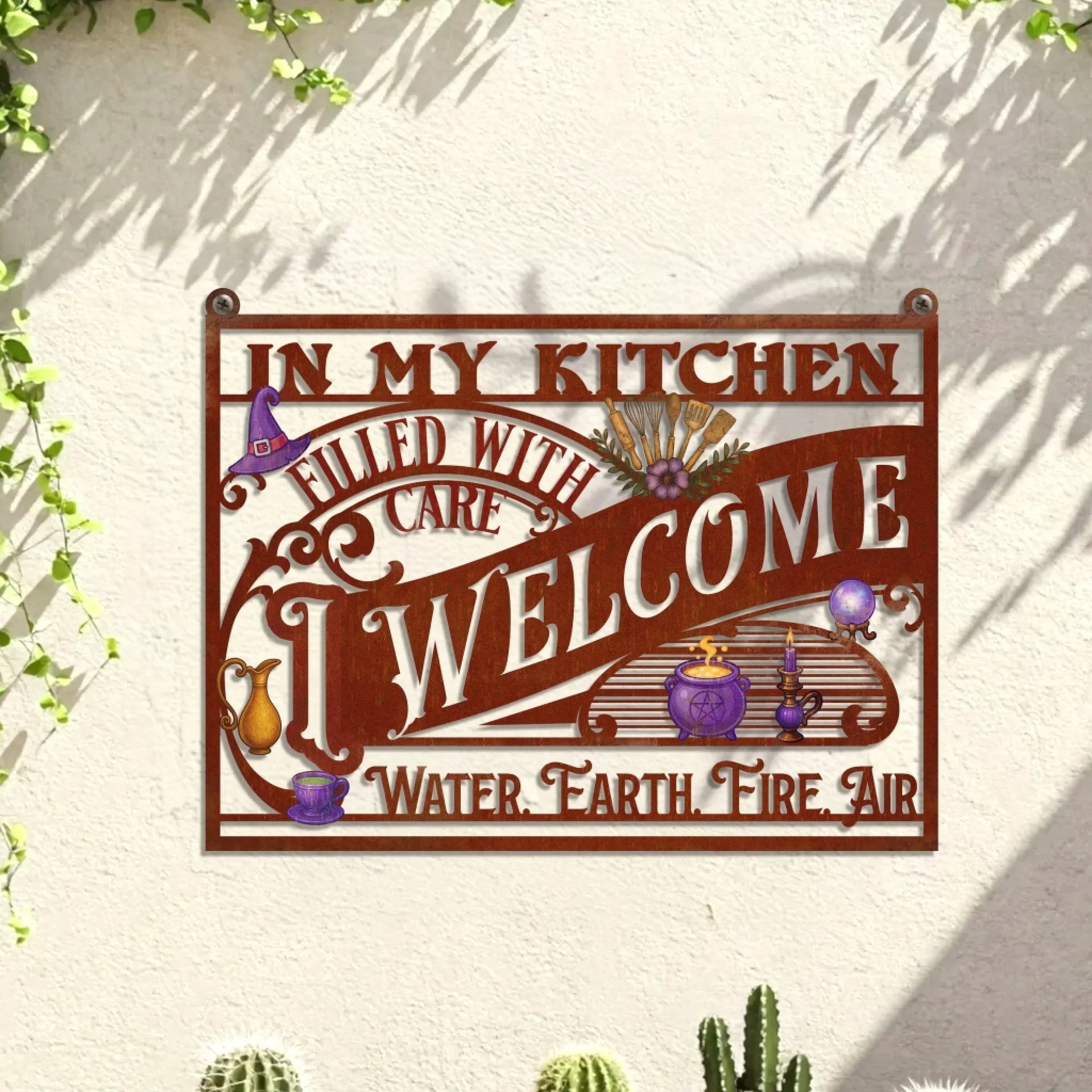 A rustic metal wall sign featuring the text In My Kitchen Filled With Care I Welcome Water Earth Fire Air with witchy-themed illustrations.