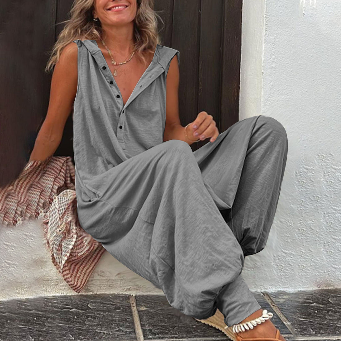 Model wearing a sleeveless hooded relaxed-fit jumpsuit in gray, showing button-front detail and tapered leg.