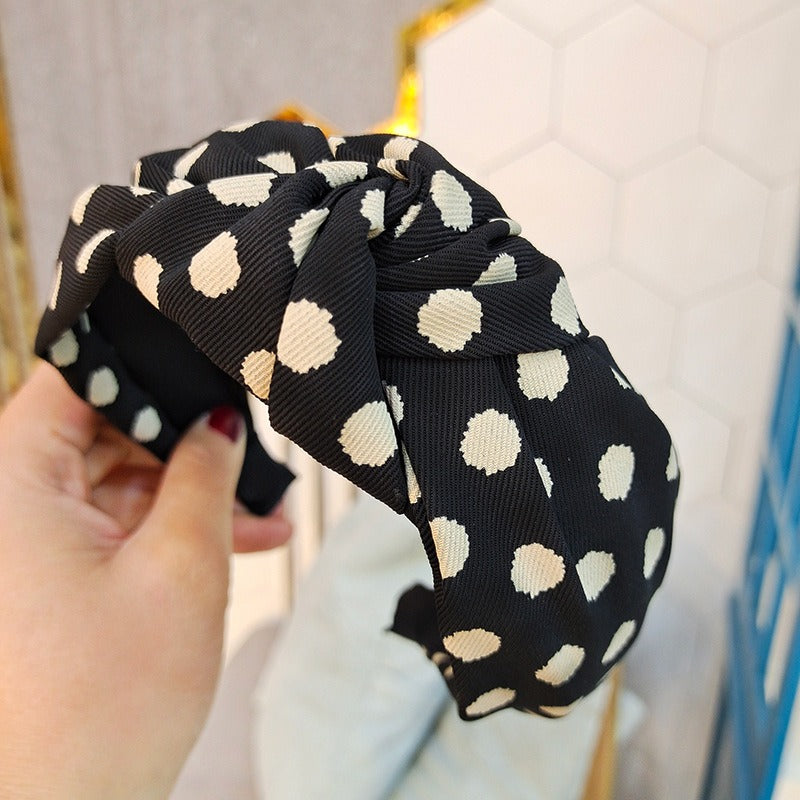 Two wide fabric polka dot headbands, one black with white dots and one light pink with black dots, shown held by a hand.