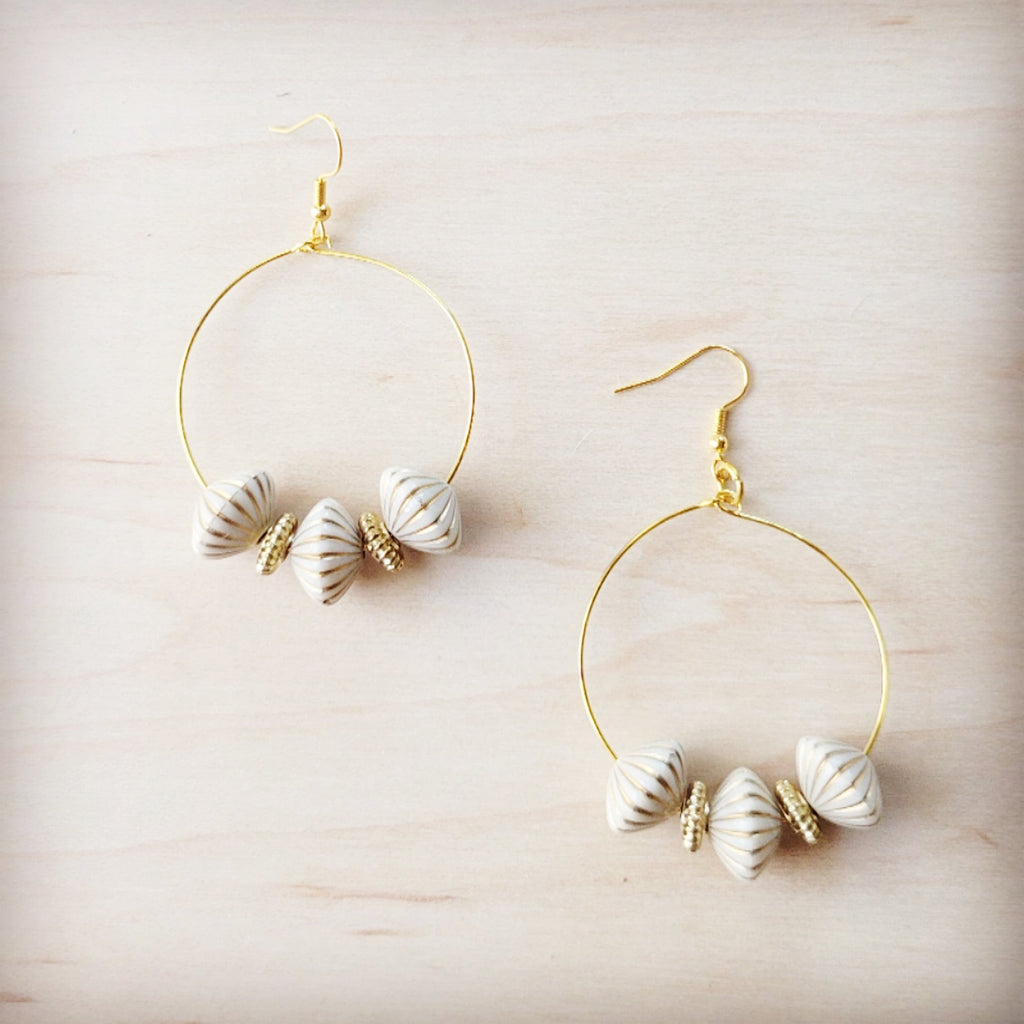A pair of gold-tone hoop earrings featuring white ridged shell beads and small gold spacers on a light wood background.