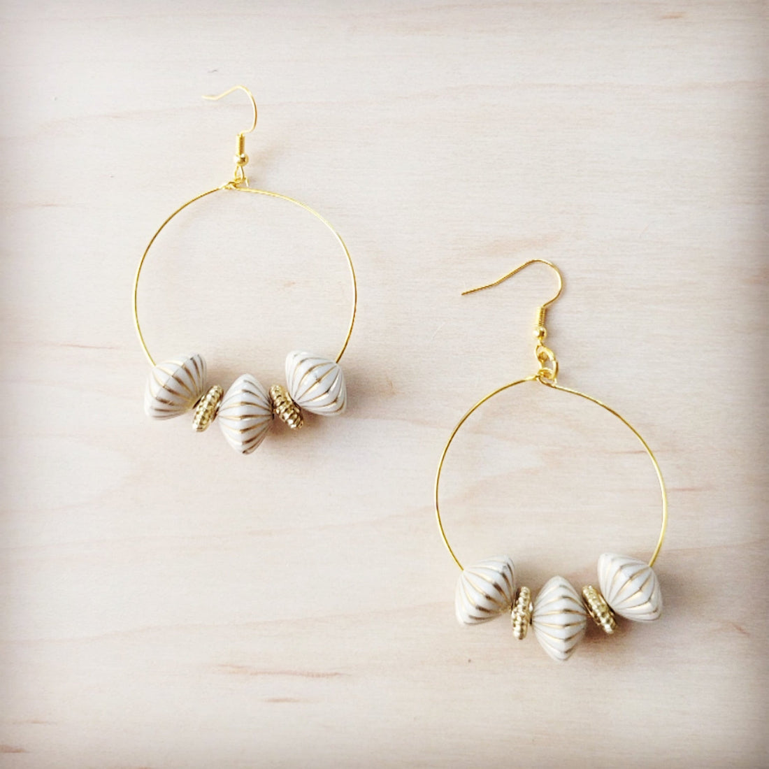 A pair of gold-tone hoop earrings featuring white ridged shell beads and small gold spacers on a light wood background.
