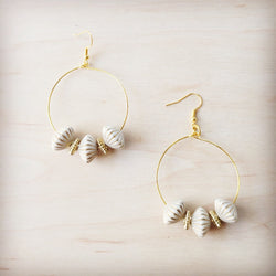 A pair of gold-tone hoop earrings featuring white ridged shell beads and small gold spacers on a light wood background.