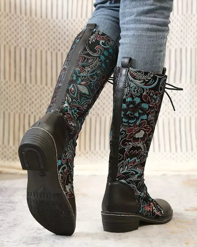 Black knee-high boots with floral pattern worn by a person indoors.