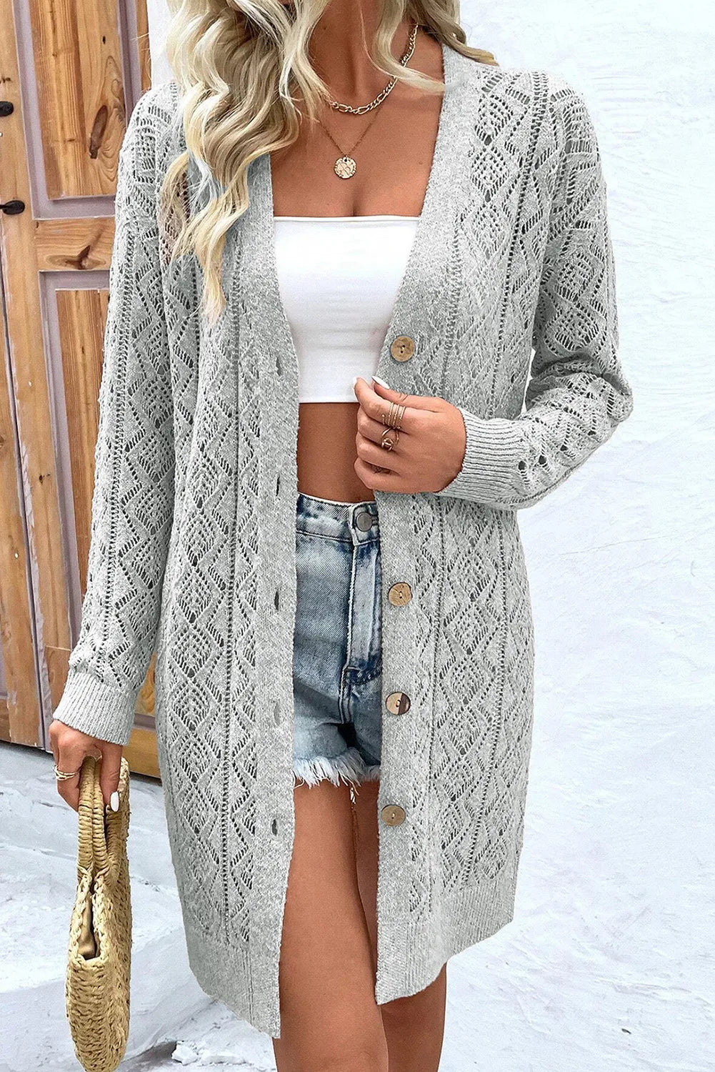 Model wearing a grey long sleeve openwork button down cardigan over a white tank top and denim shorts.