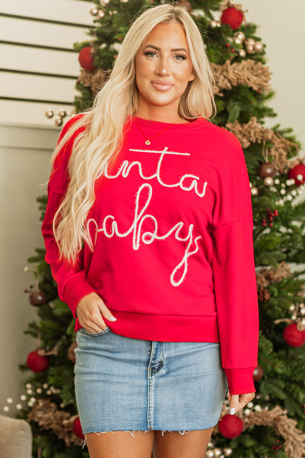 A woman wearing a vibrant red oversized sweatshirt with Santa Baby embroidered text, styled with a denim mini skirt.