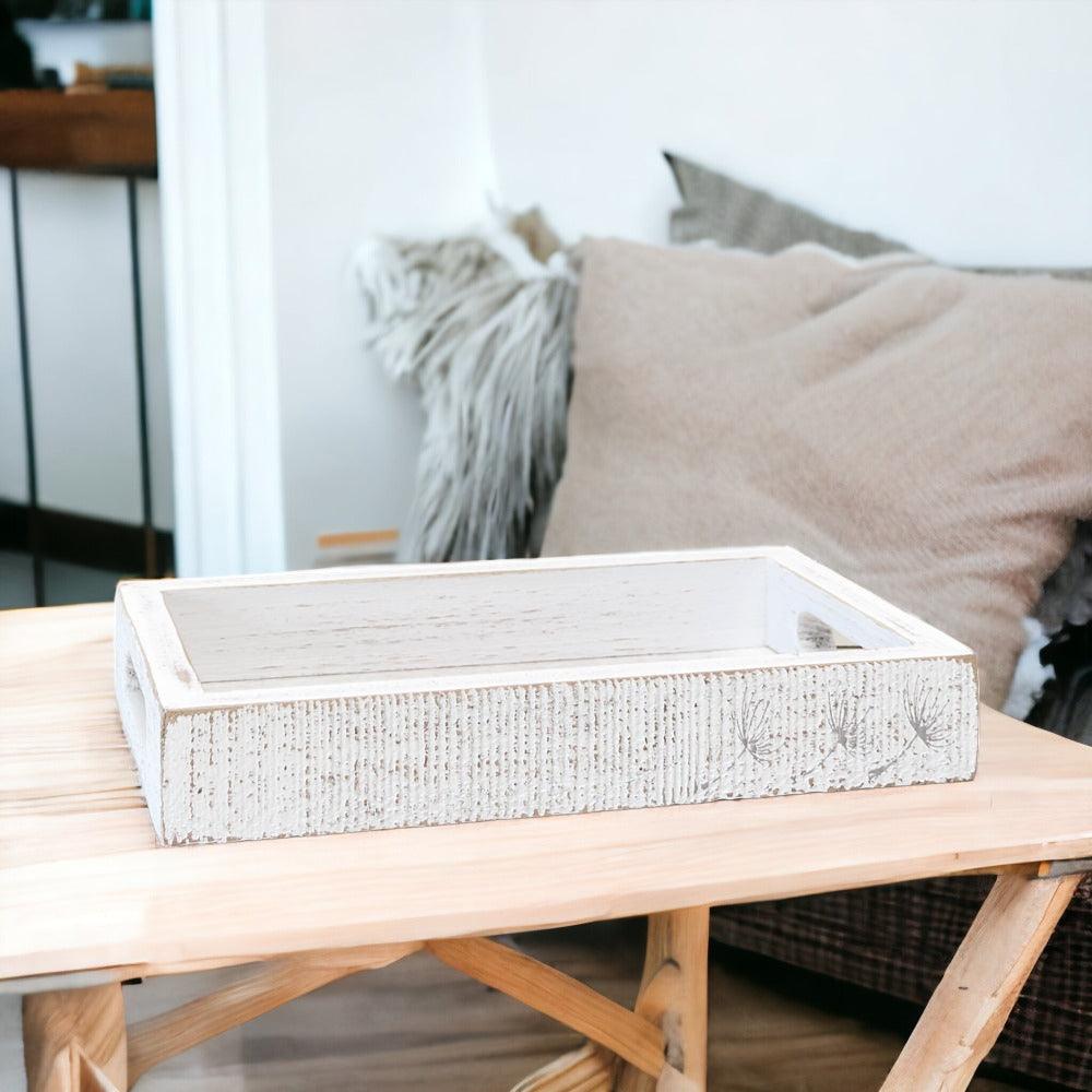 A white wash wooden decorative tray with a subtle dandelion pattern, showcased on a light wood coffee table.