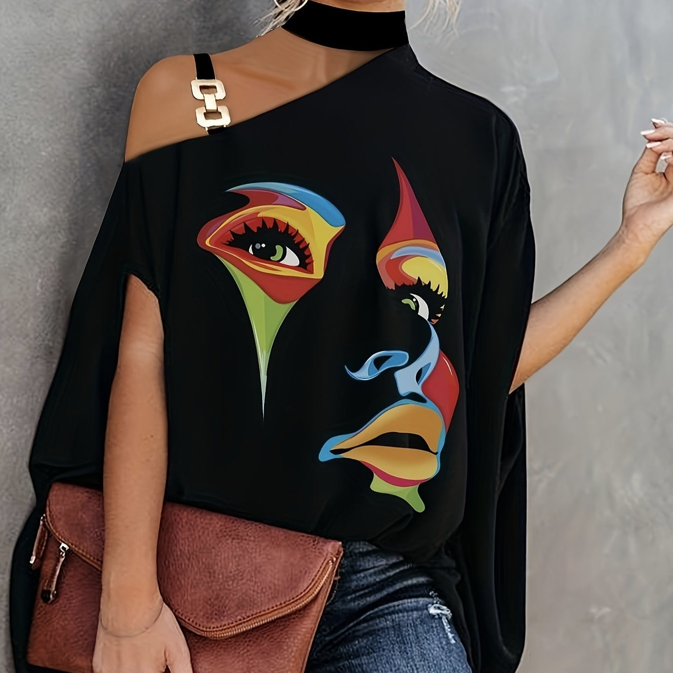 A woman wearing a black one-shoulder T-shirt with a colorful abstract face print and a metal hardware strap detail.