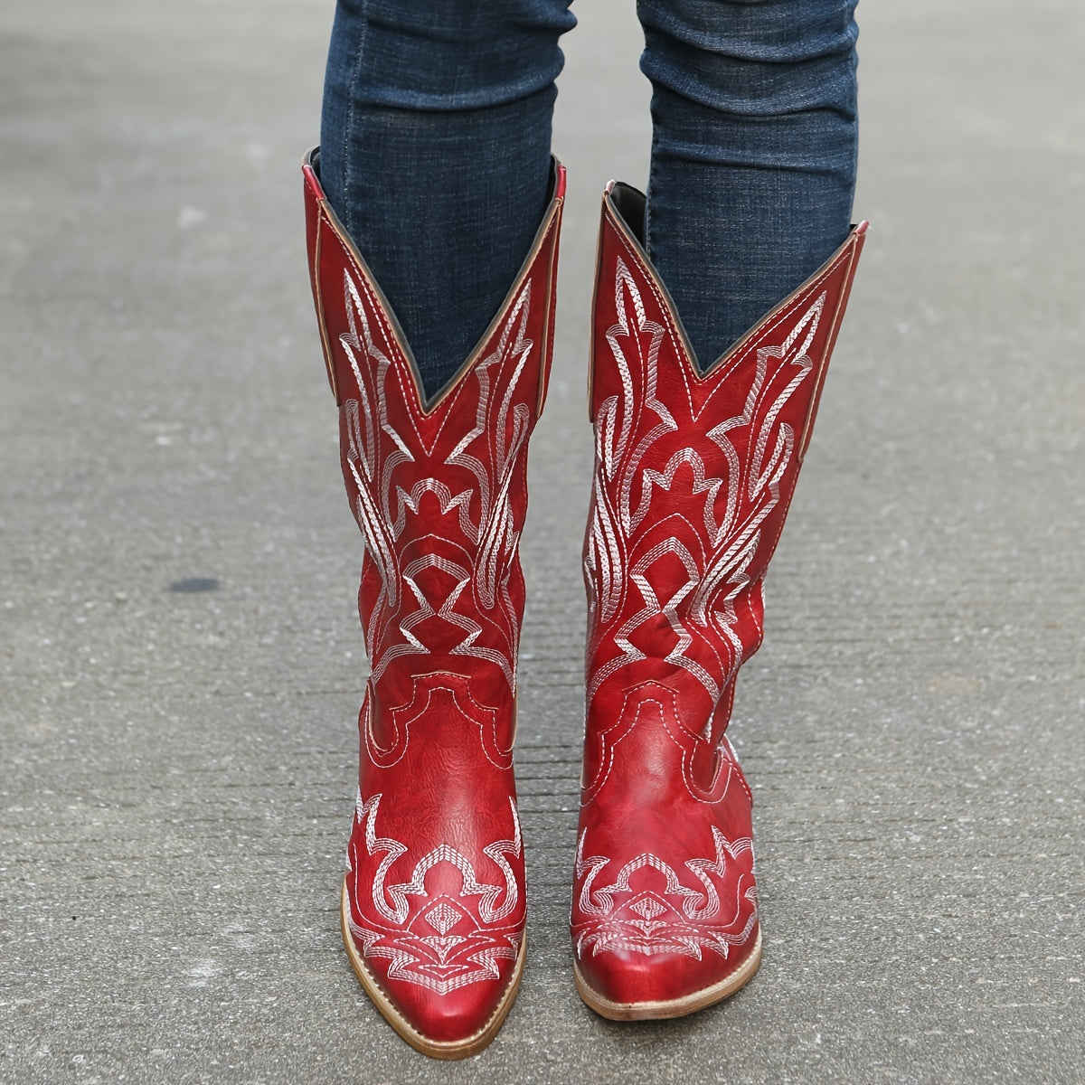 A pair of red mid-calf cowgirl boots with intricate white embroidery worn with jeans.