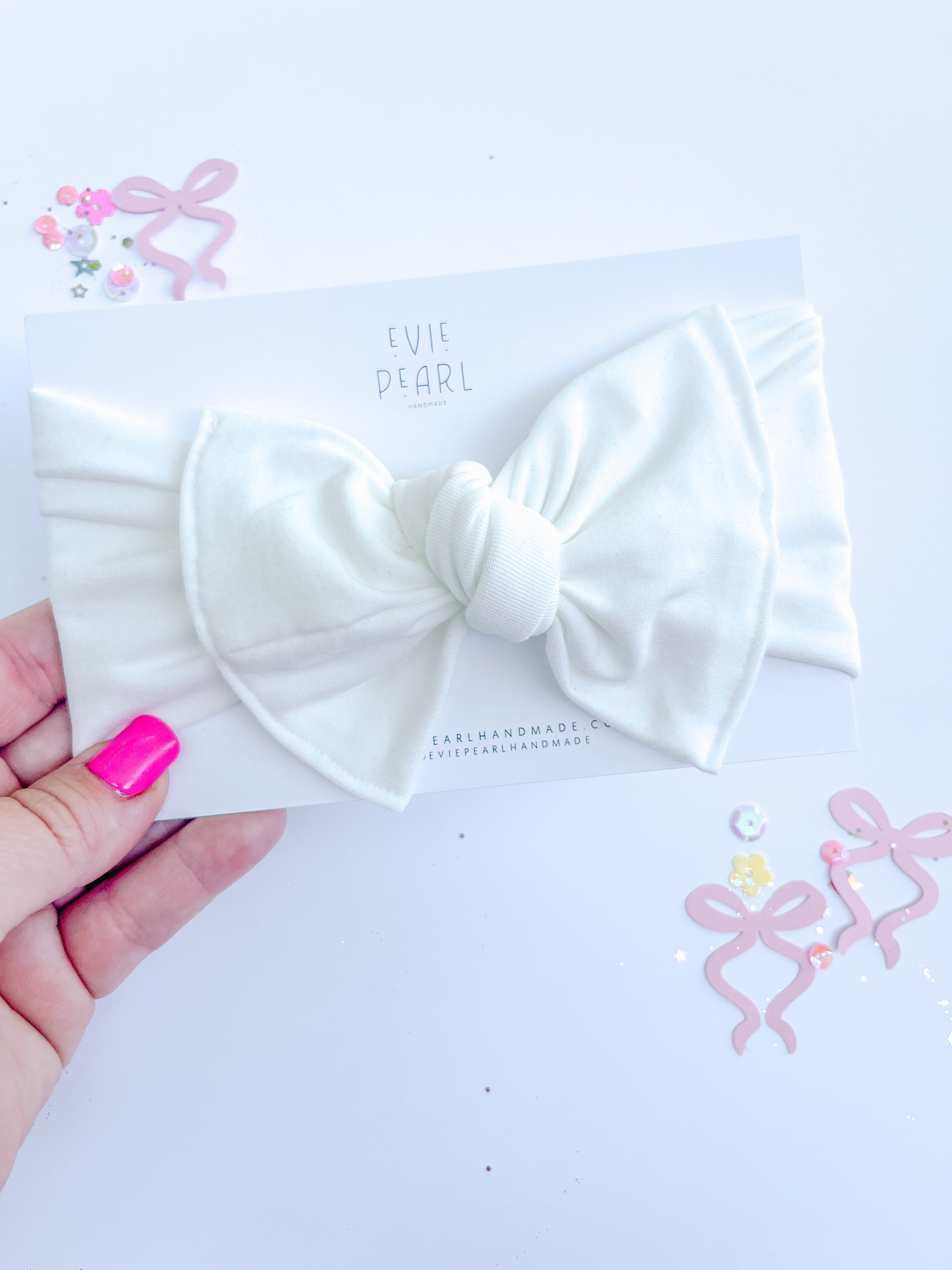 A close-up of a white, soft-knit baby turban headband featuring a large, centered bow on a product display card.