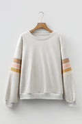 A front view of an oatmeal colored crew neck sweatshirt with horizontal pastel stripe details on the sleeves, hanging on a wooden hanger.