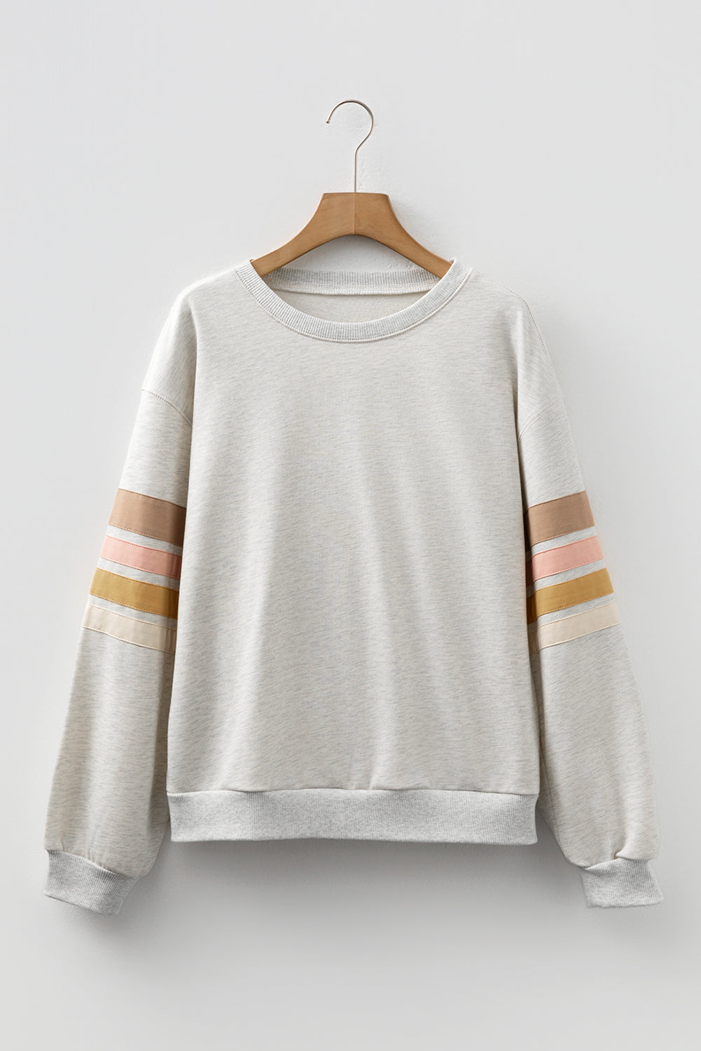 A front view of an oatmeal colored crew neck sweatshirt with horizontal pastel stripe details on the sleeves, hanging on a wooden hanger.
