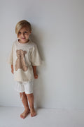 Young child wearing a beige t-shirt with a brown teddy bear print and striped shorts
