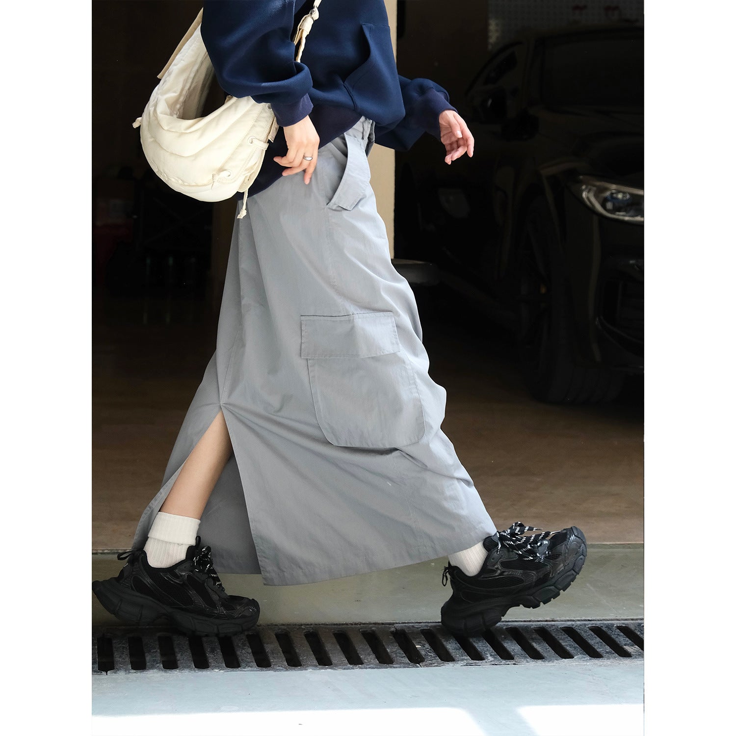 A model wears a long, gray workwear-style maxi skirt with large cargo pockets, paired with a navy hoodie and black chunky sneakers, showcasing a modern street-style look.