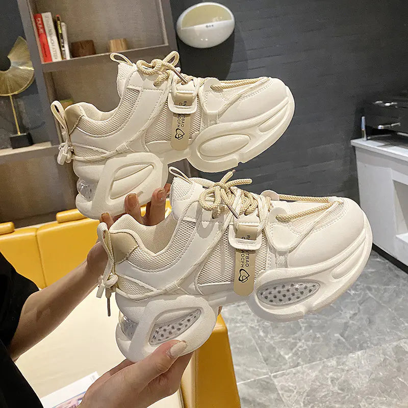A pair of trendy beige thick-soled chunky sneakers held up against a studio background.
