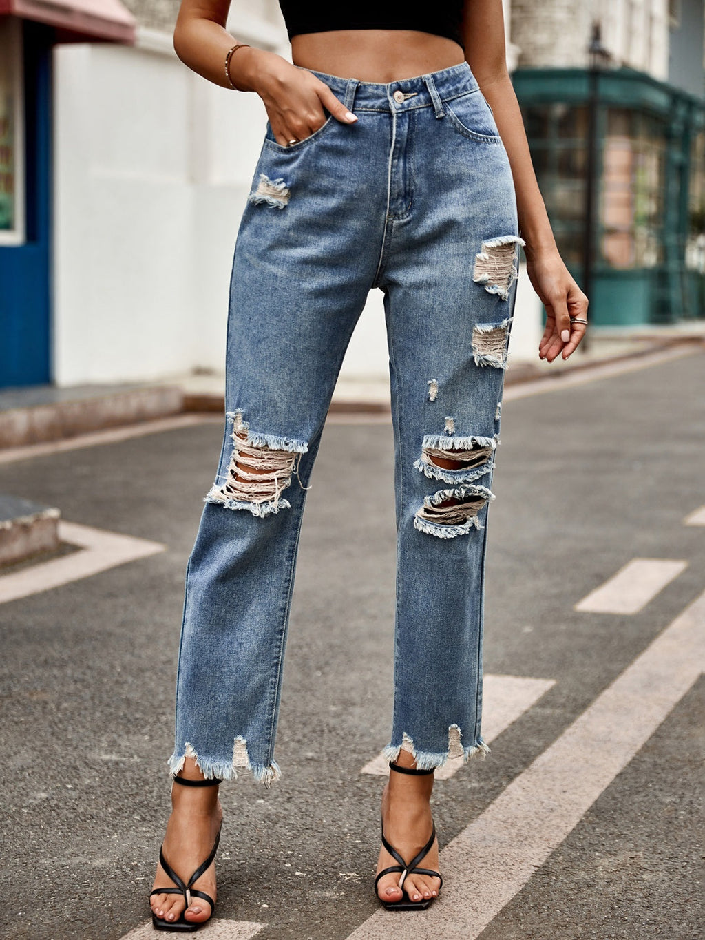 Model wearing blue denim jeans with heavy distressing on the knees and a raw frayed hem, styled with black strappy heels.