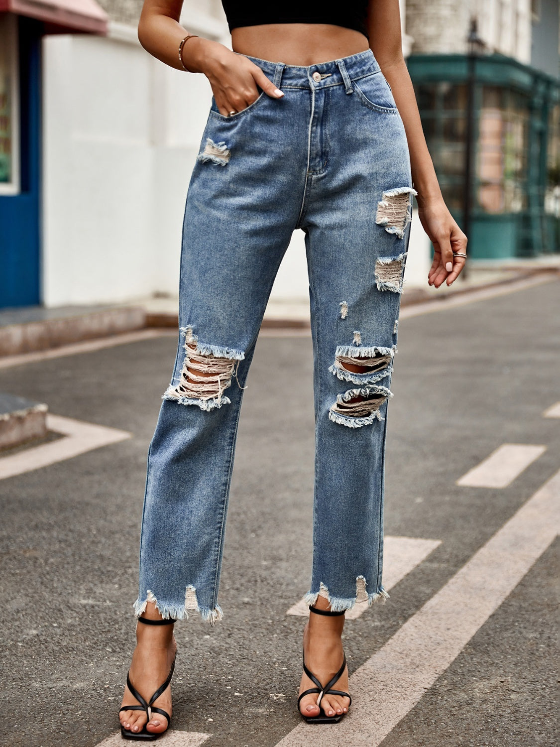 Model wearing blue denim jeans with heavy distressing on the knees and a raw frayed hem, styled with black strappy heels.
