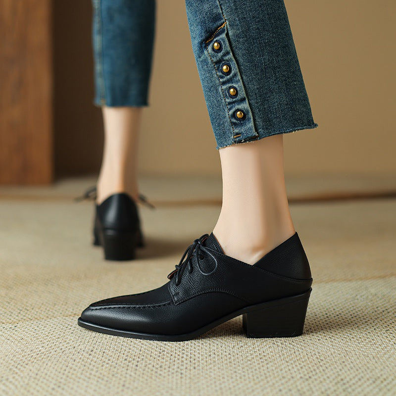 A pair of black, lace-up, pointed-toe shoes with a chunky block heel shown being held up and worn with jeans.