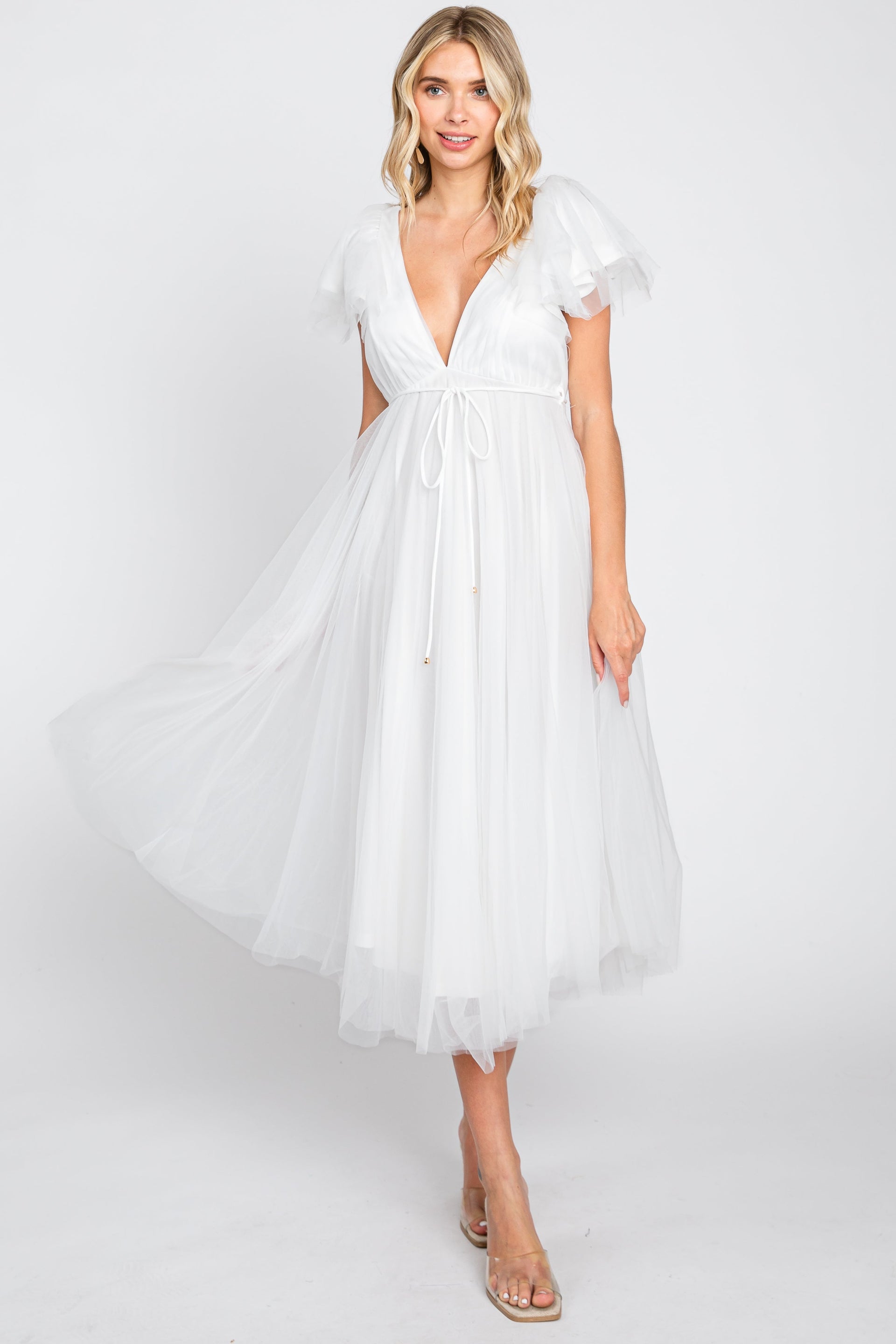A smiling woman wearing a white tulle midi dress with a v-neckline and flutter sleeves, styled with nude wedge sandals.