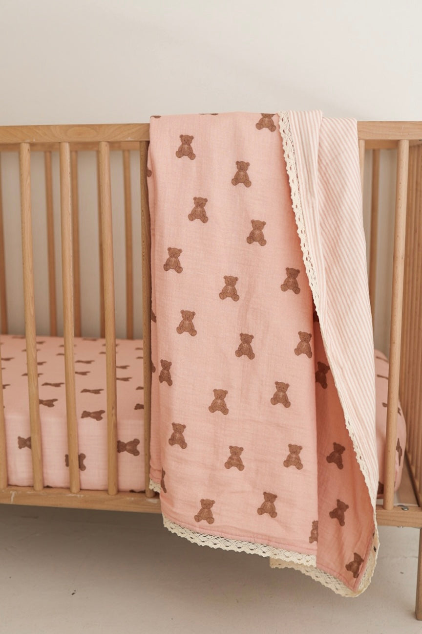 A soft pink muslin baby quilt with a brown bear print on one side and a striped pattern on the other, draped over a wooden crib.