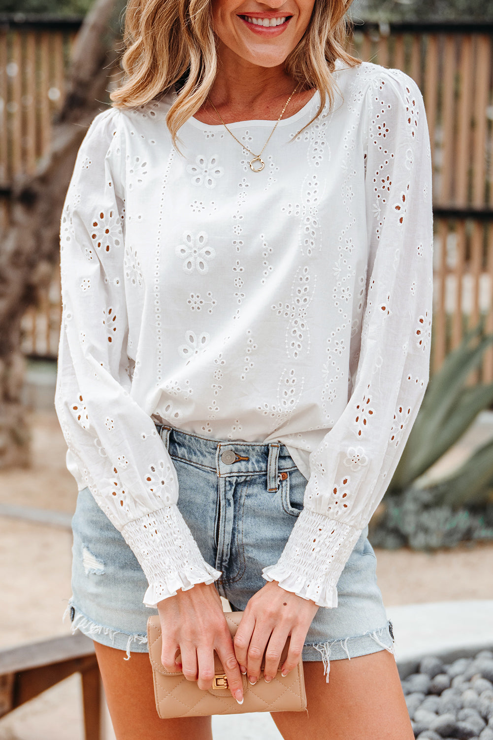 A woman wearing a white long sleeve blouse with eyelet embroidery, puff sleeves, and smocked cuffs, paired with denim shorts.