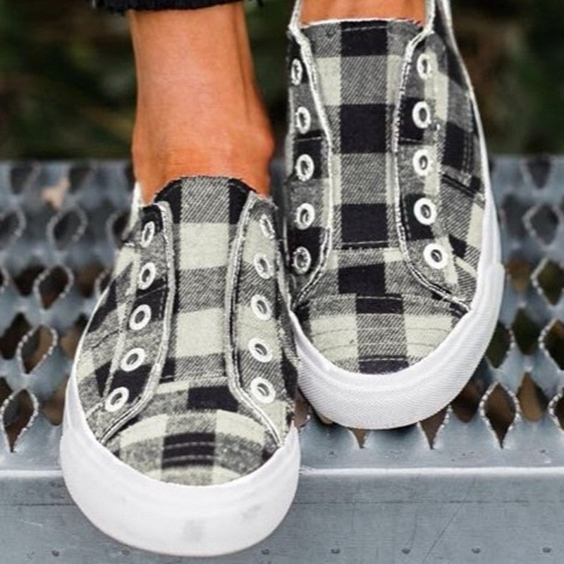 Pair of women's plaid canvas slip-on sneakers, one in black and white plaid, the other in blue and black plaid, on a metal grate.