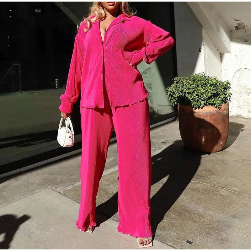 Model wearing a vibrant pink pleated two-piece set consisting of a long-sleeve button-down shirt and matching wide-leg trousers.