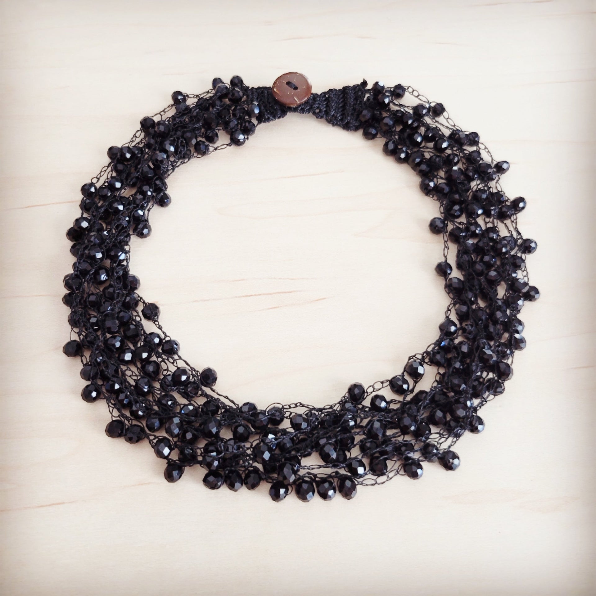 Close up of a black crystal cluster statement choker featuring a brown wooden button closure on a light wood background.