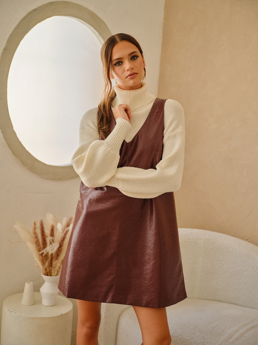 Model wearing a wine-colored faux leather shift dress layered over a cream turtleneck sweater