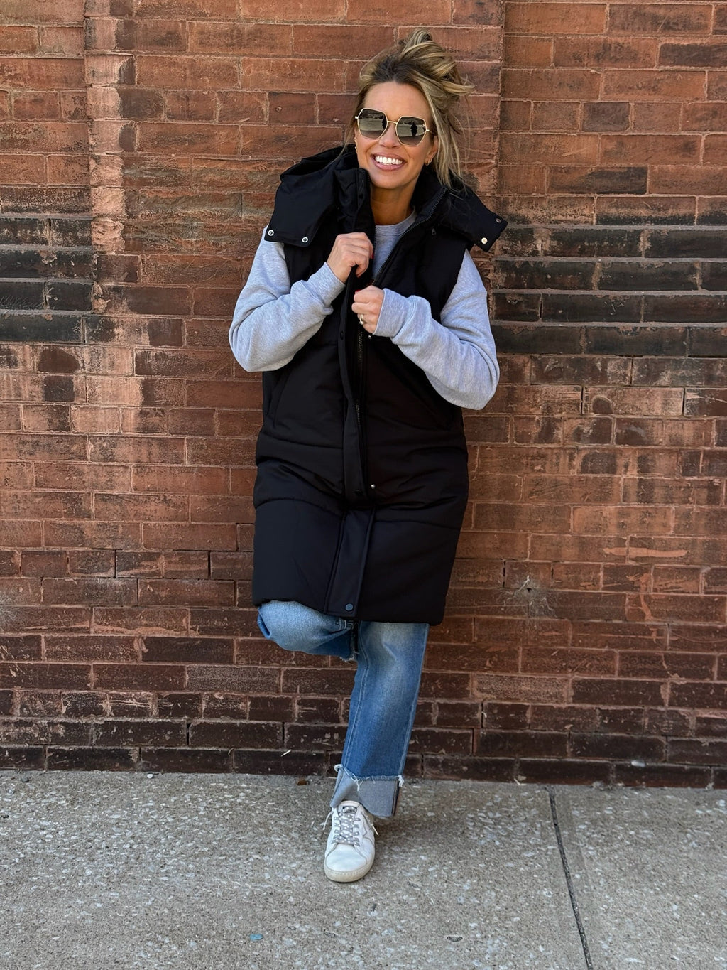A stylish long black puffer vest featuring a removable hood, front zipper and button closures, and side pockets.
