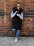 A stylish long black puffer vest featuring a removable hood, front zipper and button closures, and side pockets.