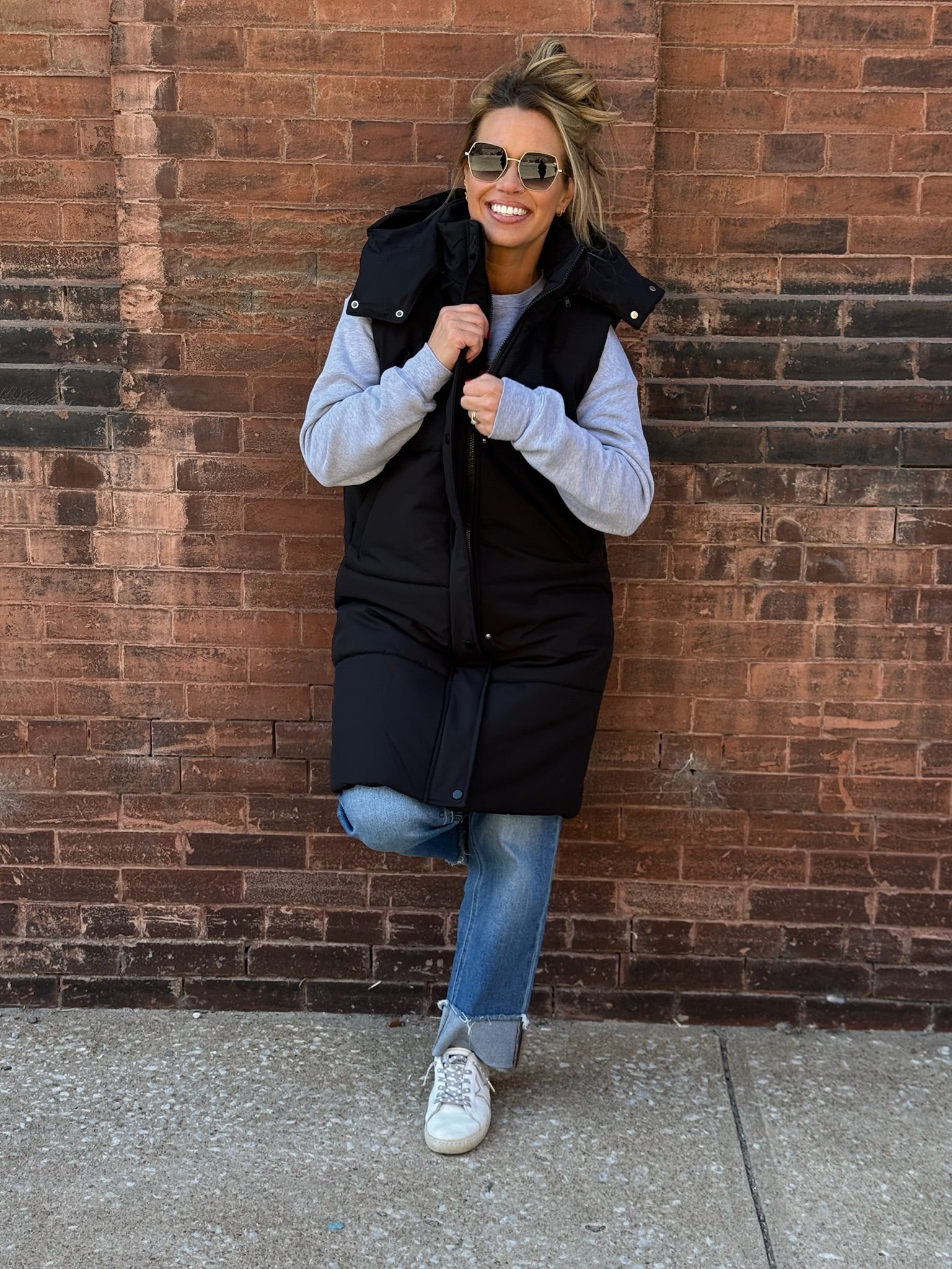 A stylish long black puffer vest featuring a removable hood, front zipper and button closures, and side pockets.