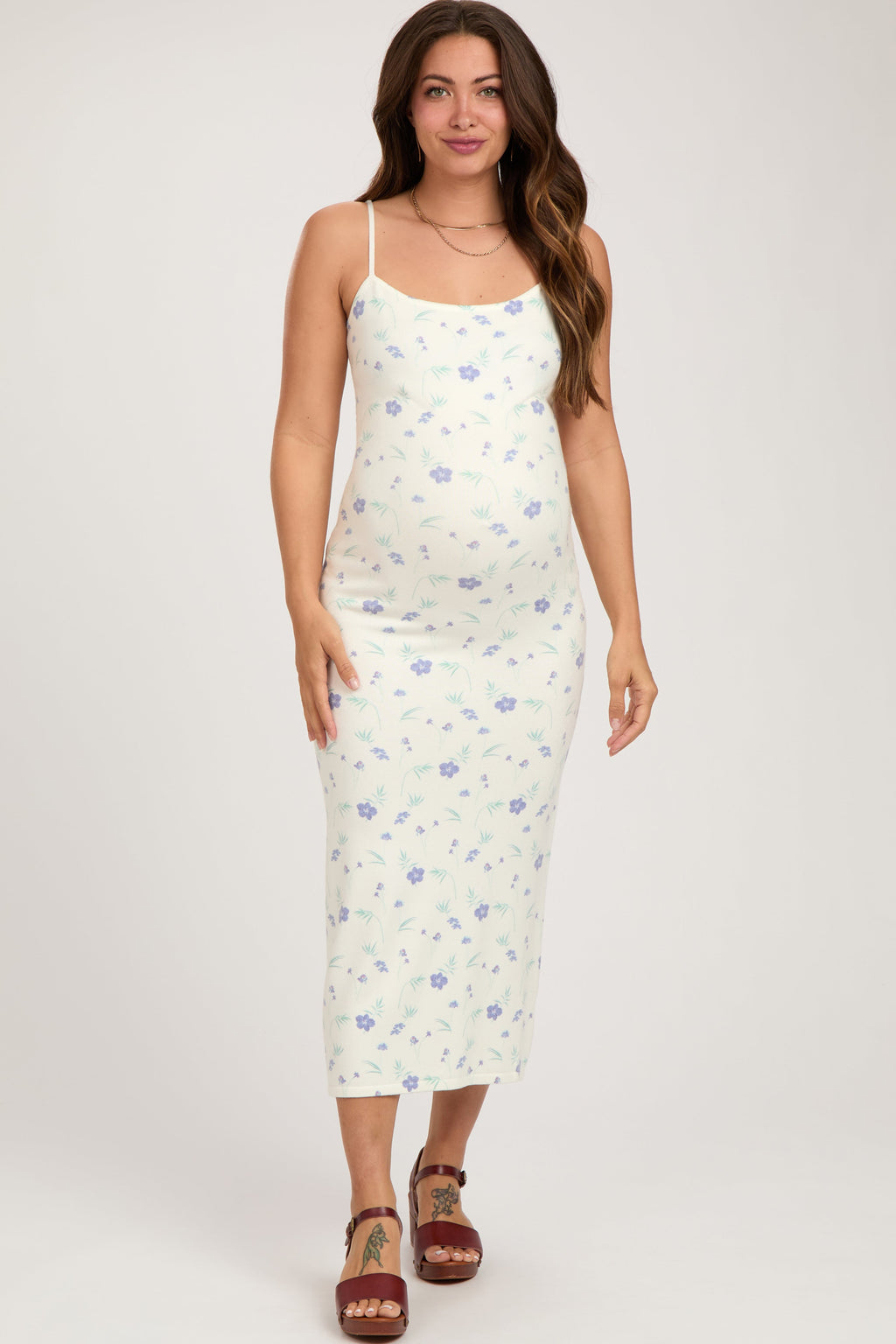 A pregnant woman wearing a white, sleeveless, knit midi dress with a floral pattern, square neckline, and adjustable straps.