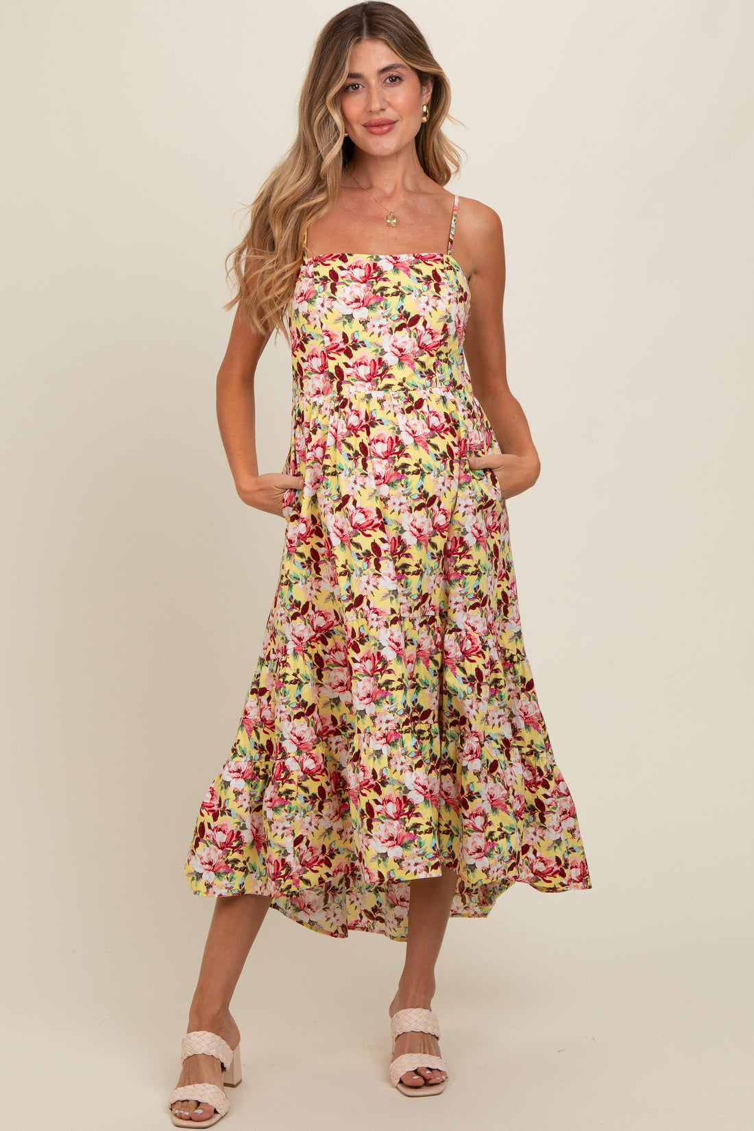 A woman wearing a yellow midi dress with a red and white floral pattern, featuring a square neckline, spaghetti straps, and a tiered hi-low hem.