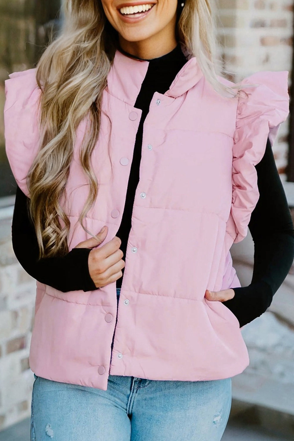 A woman wearing a soft pastel pink puffer vest with unique flutter sleeves, stand collar, and snap button closure.