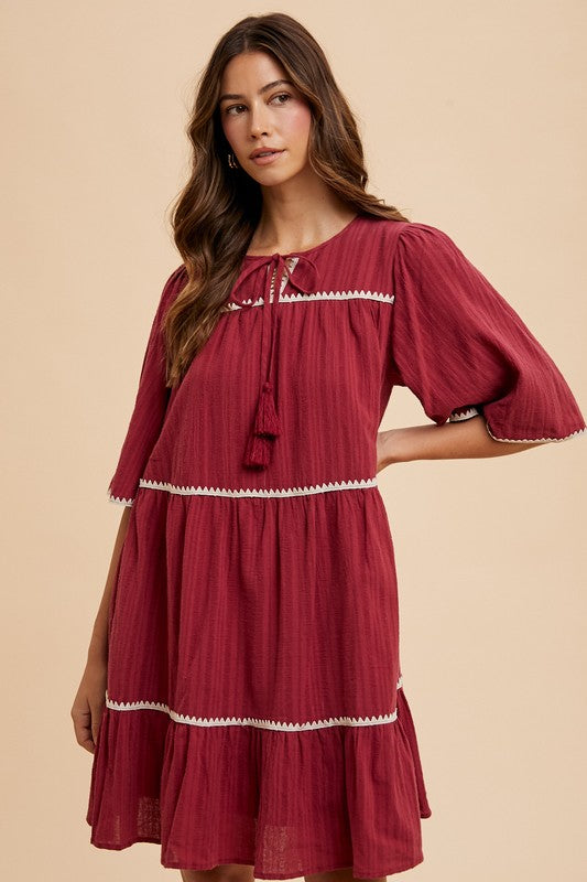 A bohemian-inspired tiered dress with tassel details and contrast trim, featuring an adjustable tie neckline.