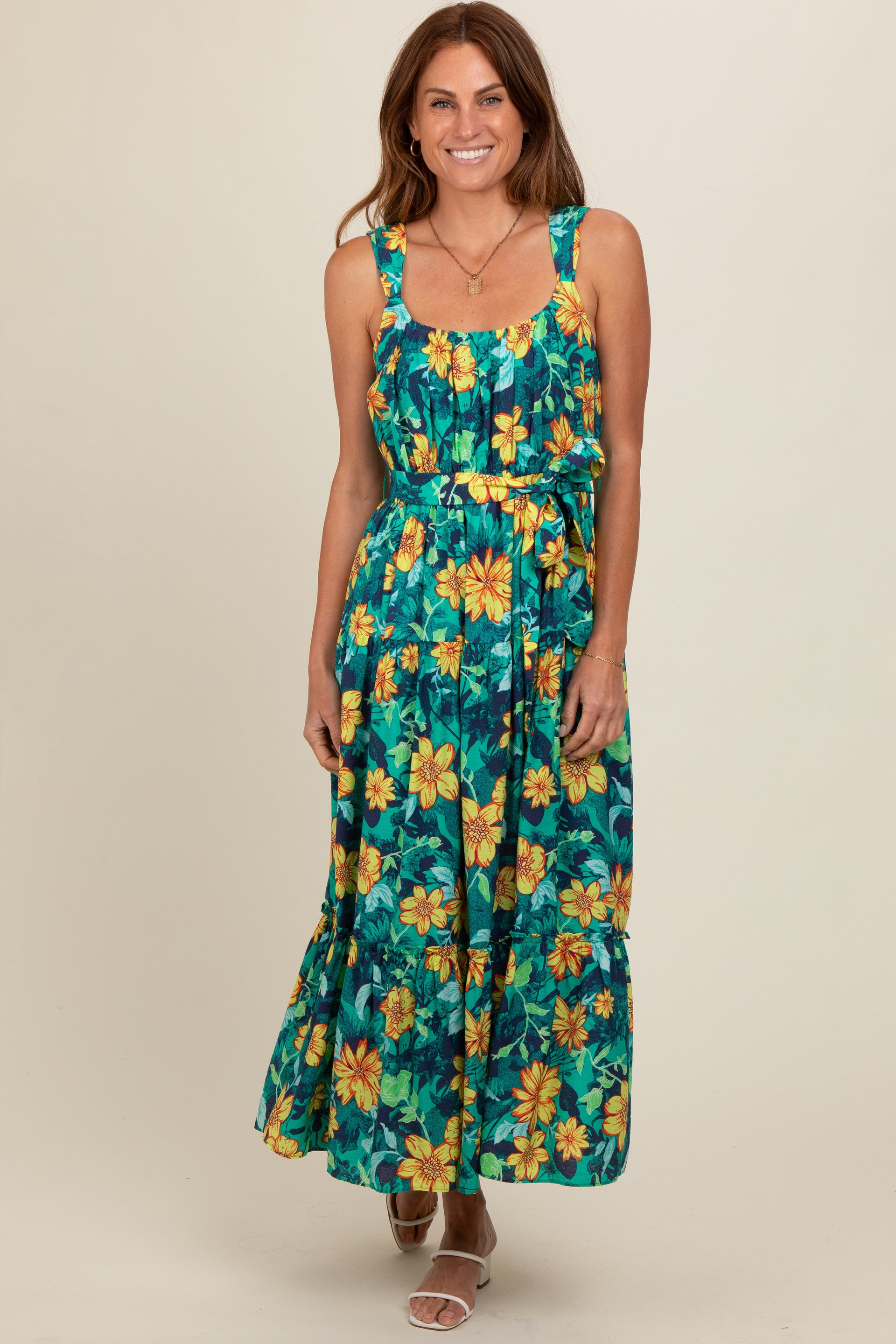 A woman wearing a yellow and teal floral tiered midi dress with a sash tie waist, standing against a neutral studio background.