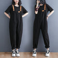 Woman wearing black Harajuku style denim overalls with tan straps and a black t-shirt, paired with white sneakers. The overalls have a relaxed fit and front patch pockets.