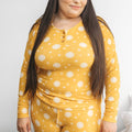 A woman wearing a yellow ribbed two-piece pajama set with a white sun and star pattern sitting on a white bed.