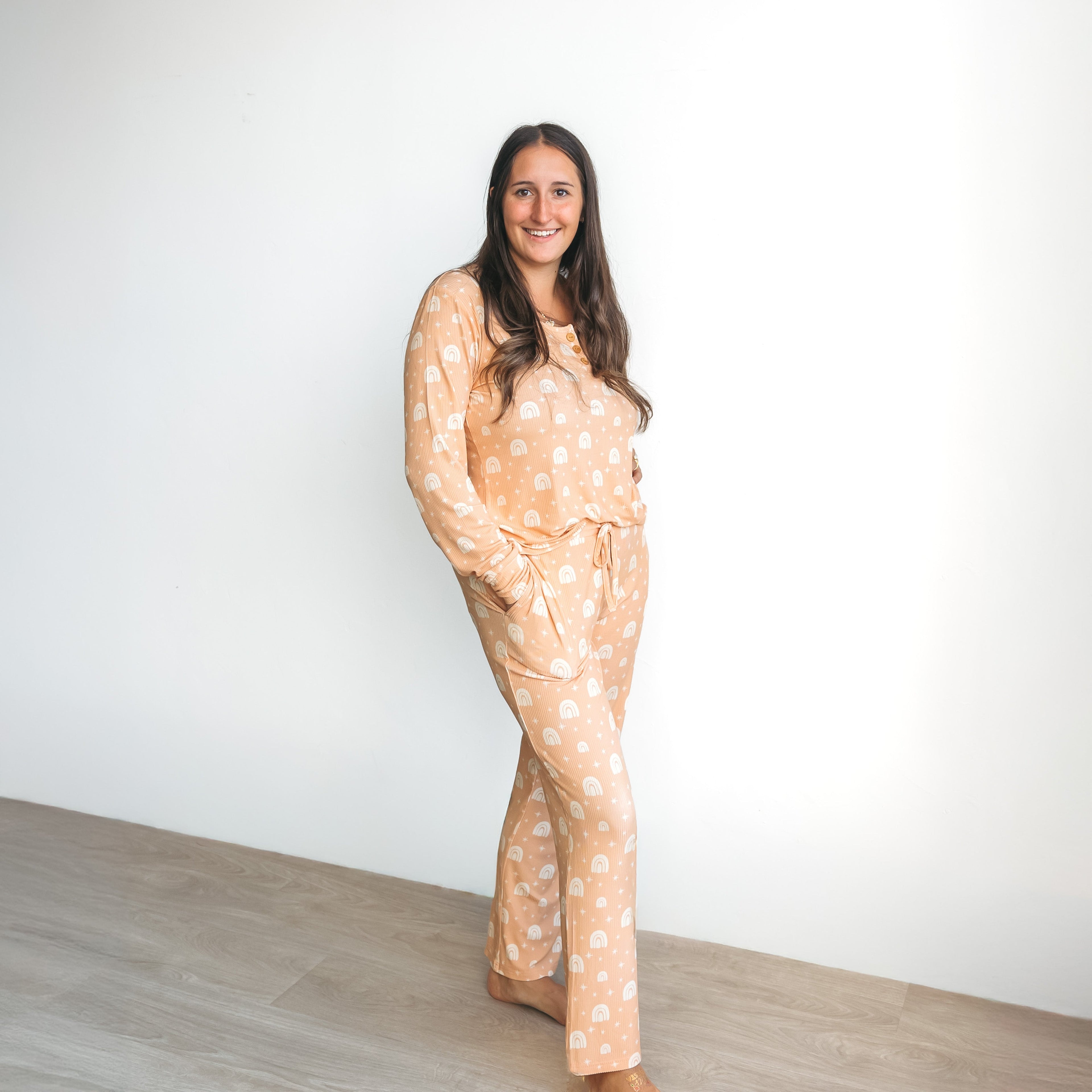 A woman wearing a peach-colored ribbed pajama set with a white rainbow pattern, featuring a button-up top and drawstring pants.