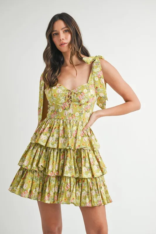 Woman wearing a romantic floral print cotton mini dress with adjustable shoulder ties in green, white, and pink.